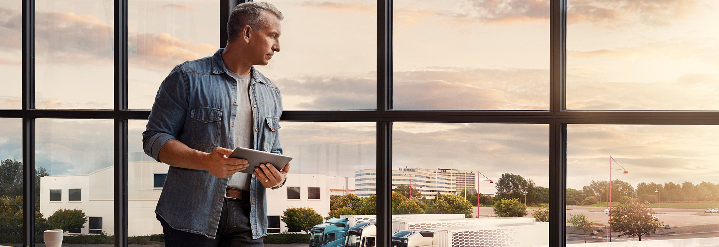 Um homem que segura um tablet junto a uma janela e observa a sua frota de camiões